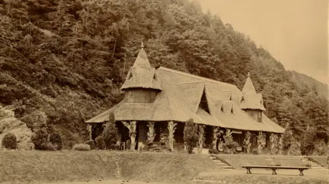 A sepia photograph showing a Swiss looking long cottage with pointy dormers raised over a lawn, on the edge of a hillside laiden with trees.