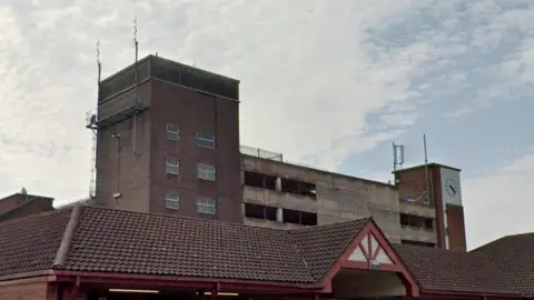 A high-rise brick multi-storey car park building is viewed behind a tiled roof with red and white gable features.