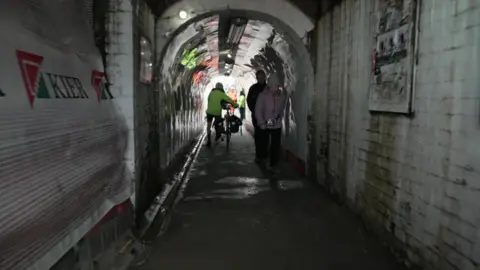 People walking through the tunnel under the Botley Road bridge. There is a person pushing a bicycle.