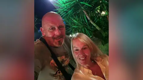 A man and a woman smile at the camera. There is a tree behind them. The man is bald and the woman has blonde hair.