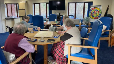 A group of elderly women sit on large blue chairs playing a board game. A colourful dart board can be seen in the background, as well as a television.