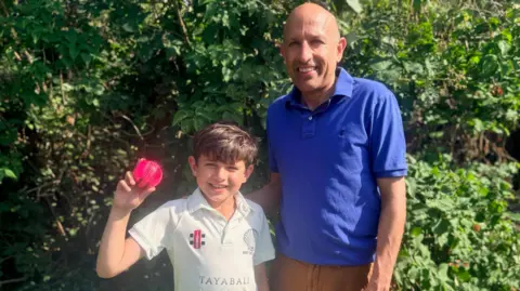 Family handout Rohan and Narain are in front of a hedge. Rohan is on the left and has short brown hair and is wearing a white cricket T-shirt. He is holding a red cricket ball near to his head. Narain is on the right and is wearing a blue polo shirt and orange trousers.