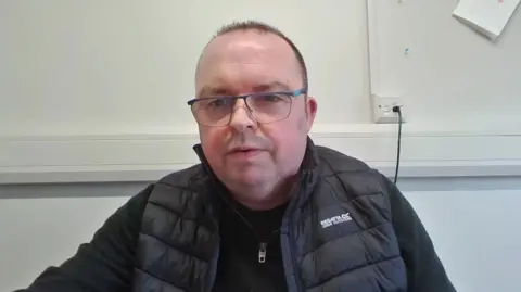 A screen grab of a video call with Sinn Féin councillor Aidan Mathers. He has very short, dair hair and glasses and is wearing a black zipped fleece under a padded black body warmer.