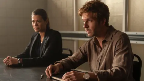 UKTV Damien Molony alongside co-star Celine Arden, sat side by side at a desk looking serious in a dark room.