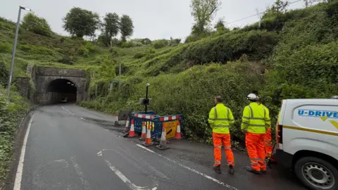 Workers in orange and yellow hi-viz stand next to a van at the entrance to Beaminster road tunnel. Overgrown embankments rise up either side of the two-way road. On the right is a small coned-off area with blue plastic barriers.