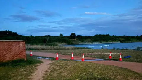 A lake with greenery around it, and orange traffic cones and a cordon in place near overflowing water.