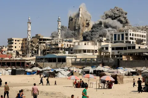 Reuters A tower block in Gaza City collapses after being hit by an Israeli air strike. Black smoke rises, and people on a beach run away