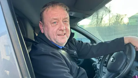 Kate Bradbrook/ BBC A man in a black Fairline branded jacket looks at the camera in the driver's seat of a van.