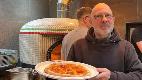 Mark Clarey with a grey beard wearing silver glasses. He is wearing a green fleece underneath a brown hoodie. He is holding a pepperoni pizza with a pizza oven behind him. 