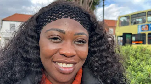 A woman with wavy black hair, held back with a wide black headband, smiles as she stands in a town square and looks into the camera. She is wearing a black coat and a red jumper. Behind her is a green hedge, a yellow double-decker bus and a white-painted building.
