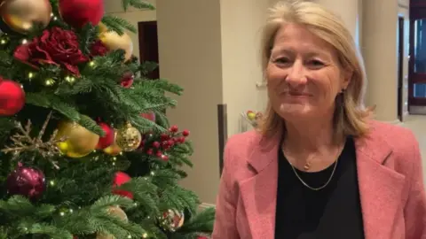 BBC Photo of a woman wearing a black shirt and pink jacket. She is sitting next to a decorated green Christmas tree.