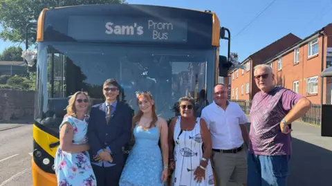 Supplied Three women and three men stand in front a bus with a yellow front. The bus reads "Sam's Prom Bus" where the route destination and number is usually displayed.