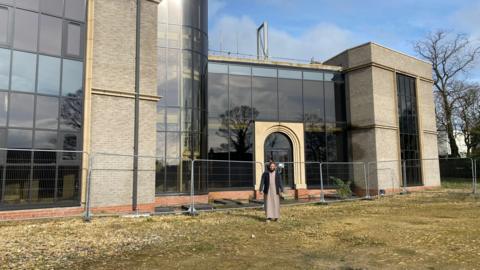 An imam standing outside a large nearly completed mosque 