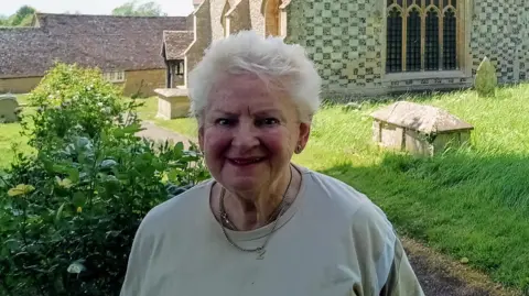 Ewelme historian Carol Sawbridge smiling for a picture in front of Ewelme Church. It is a sunny day.