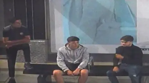 A CCTV image of three male presenting individuals. Two are sitting - one with a grey hoodie and another with a black polo neck. Another dressed in black is standing with one leg on a bench. 
