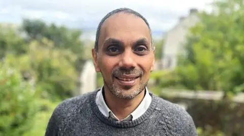 Ravinder Naga smiles at the camera standing outside with greenery behind him. He wears a grey knitted jumper and a pale grey shirt, wears his grey hair short and has a stubble beard