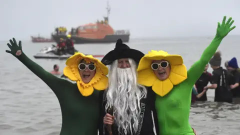 Getty Images Swimmers in fancy dress during the 52nd Boxing Day swim on December 26, 2024 in Tenby, Wales. Two people in green morph suits with daffodil hats on and wearing white glasses. Between them is a person dressed as a wizard with a long grey beard and black hat and cape and a staff.