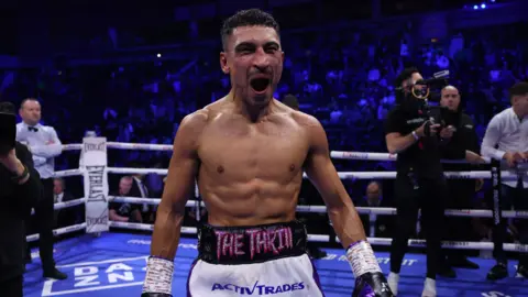 Getty Images Jordan Gill in a boxing ring after winning a bout. He has cropped black hair and a fine moustache with white trunks with 'The Thrill' embossed on the waist band.