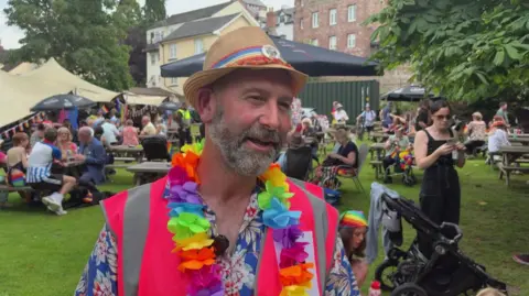 Sam is being interviewed wearing a straw hat, colourful decorations around his neck, vest and shirt. He has a short grey beard. Crowds of people can be seen in the background.