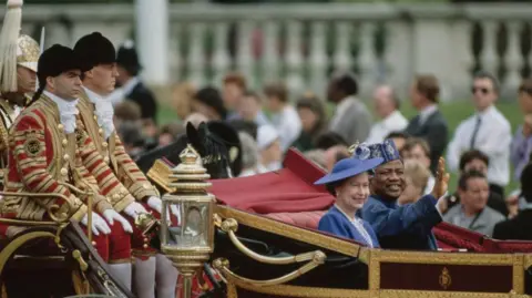 Tim Graham Photo Library via Getty Images The late Queen Elizabeth II wears a navy suit jacket, white and blue scarf, with a matching hat as she rides in a red velvet lined carriage alongside former Nigerian ruler General Ibrahim Babangida who wears a blue outfit. The background behind them is lined with on-lookers, whilst two men dressed in red and gold livery coats sit behind the carriage.