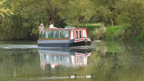 Andy Jackson A green house boat floating on the water - two people - a man and a woman can be seen standing on it. Two green and white ducks are swimming in front of it.