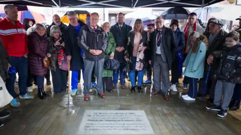 A group of people gathered outdoors under a large black canopy on a wet, rainy day. They are standing in a semicircle around a memorial stone set into the paved ground. Many of the attendees are wearing coats, scarves, and umbrellas can be seen in the background, suggesting cold or rainy weather. Some people appear to be holding programs or personal items, and a few are using walking sticks. The memorial stone on the ground is clearly visible at the center of the gathering.