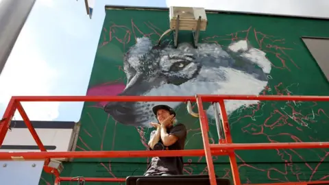 An artist is standing on red scaffolding with a large mural behind them