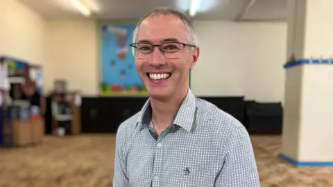 Ross Farr-Semmens is wearing a shirt and is sitting down and smiling at the camera. He is sitting inside a preschool. The background behind him is blurred. 