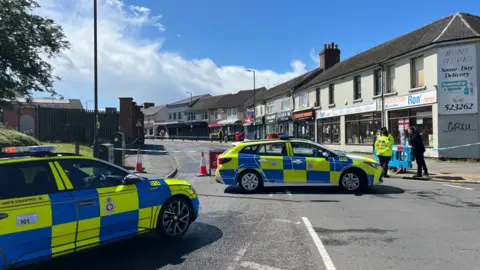 Two police cars in front of a row of shops. The road is closed, with police tape stretched across it. A number of police officers can be seen.