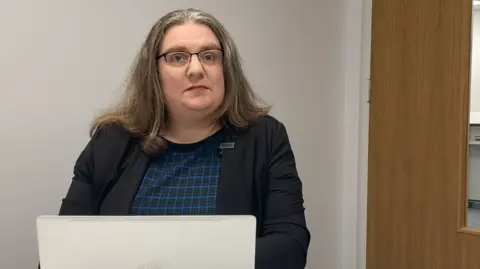 Rachel is seated at a desk in a small office room, wearing a dark blazer over a patterned top and working on an open HP laptop. A wooden door is visible to the right, and the surroundings are plain and minimal.