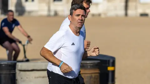 Andy Burnham wearing a white Adidas t shirt and black athletic shorts, running next to Green Park. The sun is on his face