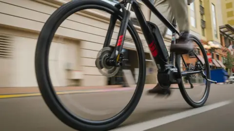 A close-up image of a black bike riding along an urban street. The bike has some red markings on it. The rider is wearing brown trousers and brown shoes.