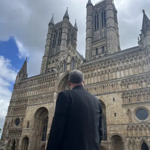 The back of a man with short grey hair and wearing a black blazer can be seen looking up at the front of a large, grand cathedral building.