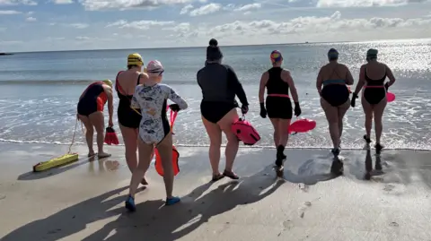 A group of women wearing swimming costumes and holding floats, walking into the sea on the beach