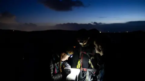 Dartmoor Plodders Three people stood in the dark with head torches lighting up a piece of paper.