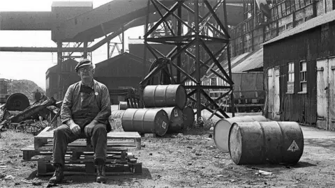 Janine Wiedel A black and white photo showing a worker sat on a pile of wooden pallets. There are a number of metal barrels lying on their side in the area. Behind is an industrial scene with factory buildings.