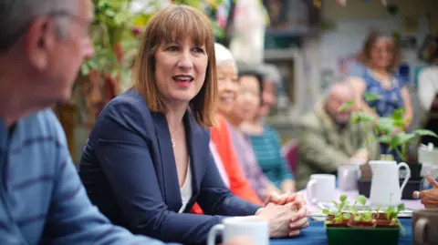 Rachel Reeves sat at a table surrounded by members of the public, with a plant pot in front of her and various cups and jugs.