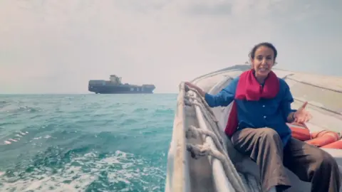 Nawal wears a scarf, shirt and trousers as she sits in a boat in the strait with a container ship seen in the background.Nawal wears a scarf, shirt and trousers as she sits in a boat on the Strait with a shipping container seen in the background