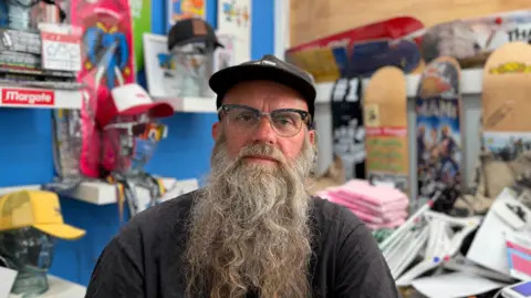 A man with a long grey beard and glasses, wearing a cap, sitting in a shop in front of a row of skateboards