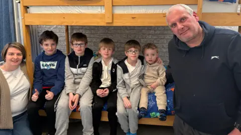 Mother Sam stands to the far left wearing a white T-shirt, cardigan and jeans. She has a short bob hairstyle. Next to her, sitting on the bottom bunk bed are her five sons all lined up together wearing tracksuits. Their father Jason is standing on the right wearing a navy hooded top. He has short grey hair.