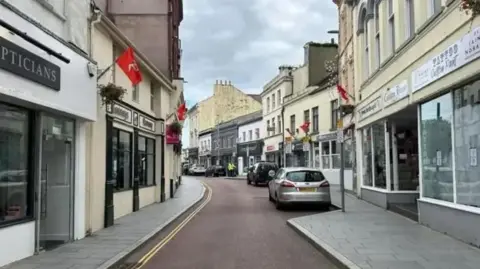 Ramsey high street, with two rows of shops including an opticians, a tattoo parlour and a pharmacy. Red Manx flags are dotted along the street above the shops. The tarmac of the road separating the rows is red, and cars are parked along the right-hand side of the street.