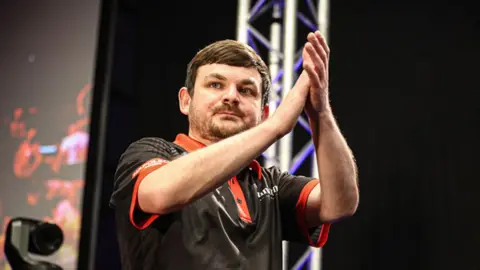 PDC Europe A man with dark hair and wearing a mainly dark top has his hands together.