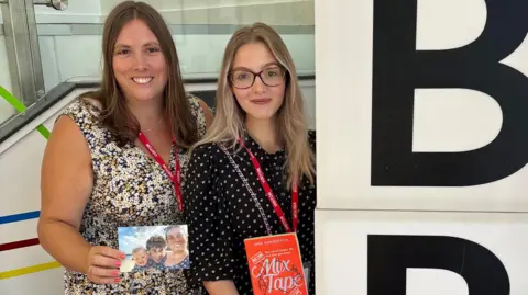 BBC Nicola Hellem wears a flowery white, blue and yellow dress and smiles as she holds a photograph depicting herself with her daughter and husband on a sunny day in Looe, while next to her stands a smiling Emily Morse wearing a black and white spotty dress and a book called Mix Tape. The first letters of the BBC sign can be seen to their right.