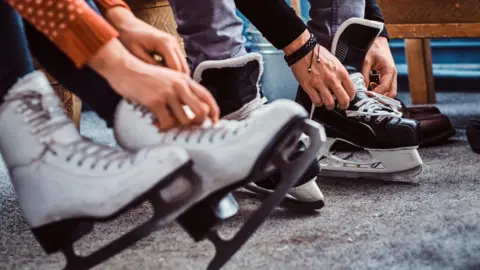 Row of ice skates on people's feet with hands doing up the laces.