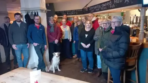 Nine men, six women and a white and brown dog stand in a pub. They are dressed in winter clothes. There is a large white column, blackboards above the bar, a large wooden table and a wooden bar stool.