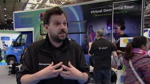 A man with dark hair and facial hair dressed in a black t-shirt and zip jumper with a white logo on it that reads "Training 2 Care". He is standing in an exhibition hall, talking and gesturing with his hands