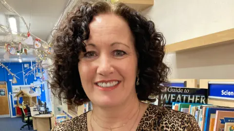 A woman with dark curly hair in a school library with books on shelves behind her and some Christmas decorations