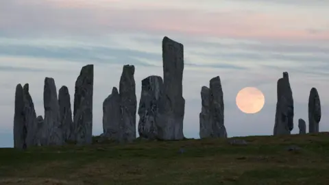 A ring of tall, grey stones in an area of short grass. The sky is a pale blue with thin cloud and the Moon is low in the sky and has a pink-orange hue.