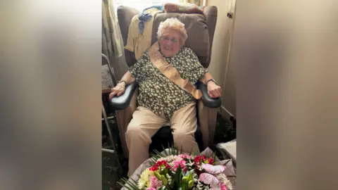 Family photo Phyliss Marshall sat inside her home with a big bouquet of flowers in front of her. She is wearing a green blouse with white spots across it. She is smiling and has a birthday sash across her.