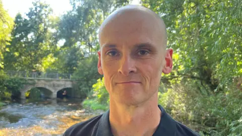 Richard Daniel/BBC James Heywood is wearing a dark polo shirt and standing in front of a bridge and stream.